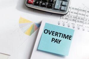 A sticky note that says “overtime pay” sits on top of a cluttered desk.