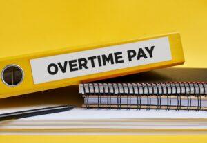 A wooden block with “Overtime Pay” written on it next to a clock and on top of money, with someone nearby wondering whether unpaid overtime is legal in California